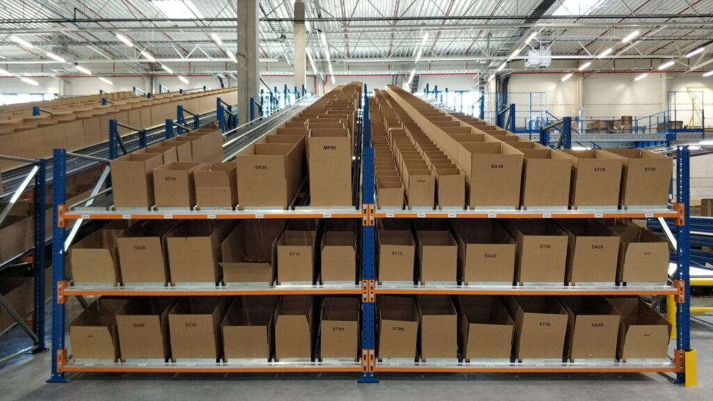 System regałów przepływowych z kartonowymi pojemnikami w nowoczesnym centrum logistycznym. Każdy pojemnik oznaczony jest unikalnym numerem, co ułatwia identyfikację i kompletację zamówień. Widoczne tory rolkowe umożliwiają grawitacyjne przesuwanie opakowań, wspierając efektywny przepływ towarów. Infrastruktura ta stanowi przykład innowacyjnych rozwiązań zwiększających wydajność operacji magazynowych.