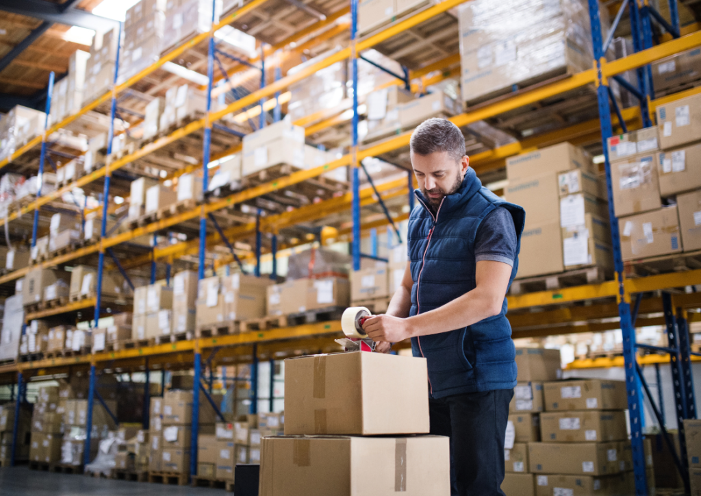 Pracownik magazynu pakujący kartony w otoczeniu regałów wysokiego składowania – nowoczesne i zorganizowane środowisko pracy