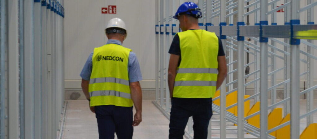 Kierownik magazynu omawiający zakres obowiązków z pracownikiem w nowoczesnym magazynie Nedcon, co ilustruje zarządzanie personelem i procesami.