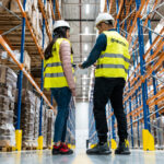 Dwóch specjalistów Nedcon (kobieta i mężczyzna) w kamizelkach odblaskowych i kaskach ochronnych stoi w wąskim korytarzu wysokiego regału paletowego, rozmawiając i analizując dokumenty. W tle widoczne rzędy magazynowanych produktów.